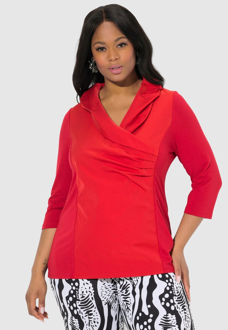 Rotes Longsleeve-Oberteil mit drapiertem Kragen, das über gefaltete Details verfügt. Kombiniert mit schwarz-weißen gemusterten Hosen. Glatte Textur.