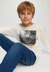 Biała koszulka z długim rękawem z graficznym nadrukiem przedstawiającym kosz do koszykówki, połączona z ciemnoniebieskimi dżinsami. Gładki materiał, luźny krój.