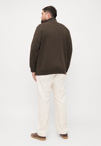 Sweatshirt marron foncé avec col montant, poignets côtelés, associé à un pantalon couleur crème et des chaussures marron à rayures. Design minimaliste.
