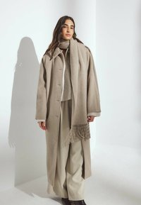Bežový vlnený kabát s golierovým dizajnom, gombíkovým zapínaním a veľkým strihom, v kombinácii s ladným šálom. Model má na sebe krémové široké nohavice.