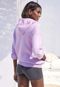 Rosa Kapuzenshirt mit dem Aufdruck "SAND" auf dem Rücken, kombiniert mit dunkelgrauen Shorts. Weicher Stoff, lockere Passform, kurze Ärmel, minimalistisches Design.