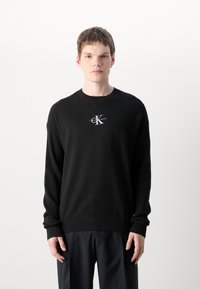 Black knit sweater with ribbed texture, crew neckline, and long sleeves. Features a white embroidered logo on the chest.