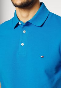 Mu muž v jasně modrém polo tričku s třemi bílými knoflíky a malým červeno-bílo-modrým logem na levé hrudi.