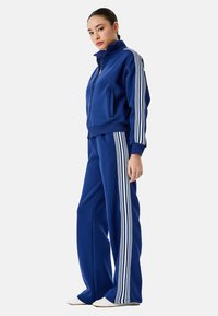 Blauer Trainingsanzug aus weichem Stoff, bestehend aus einer Jacke mit hohem Kragen und Frontreißverschluss sowie weißen Streifen an den Ärmeln und Seiten der Hose.