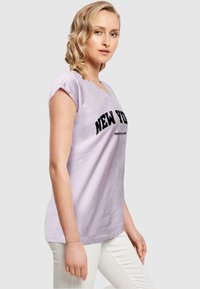 Lyserød kortærmet t-shirt med v-udskæring og sort "NEW YORK" grafik trykt på forsiden. Kombineret med hvide bukser.