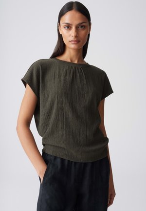 Femme aux longs cheveux foncés portant un haut texturé olive à manches courtes et un pantalon noir, debout avec une main dans la poche.