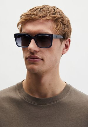 Jonge man met kort, krullend blond haar, die donkere, rechthoekige zonnebril draagt en een bruin shirt met ronde hals, tegen een effen achtergrond.