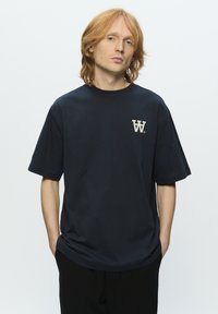 Mørkeblå bomulds t-shirt med korte ærmer, med et beige-logo på venstre brystområde. Løs pasform og ensartet tekstur.