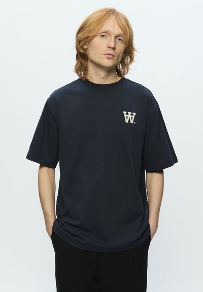 T-shirt en coton bleu marine à manches courtes, arborant un logo beige sur la poitrine gauche. Coupe décontractée et texture solide.