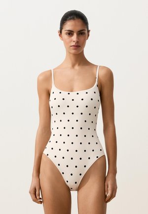 Nő fehér, fekete pöttyös egyrészes fürdőruhában, vékony vállpántokkal, világos, egyszínű háttér előtt állva.