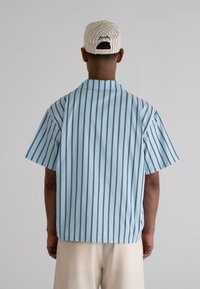Light blue short-sleeve shirt with vertical navy stripes, collared design, and a straight cut. Worn with a beige striped baseball cap.