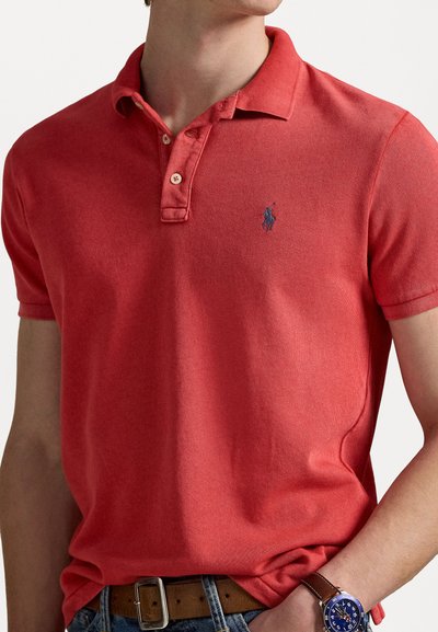 Rød polo-skjorte laget av teksturert stoff, med knappekrage, korte ermer, og en liten blå brodert logo på brystet.