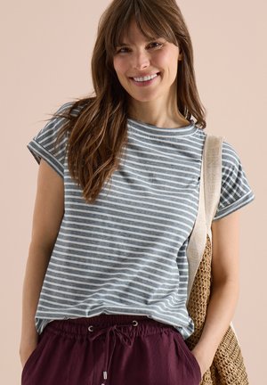 Femme souriante portant un t-shirt rayé gris et blanc, un pantalon bordeaux à cordon et un sac tissé sur une épaule.