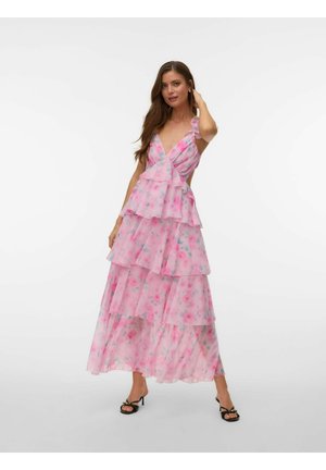 Kvinne iført rosa blomstrete lagen lang maxikjole med volanger og svarte sandaler med hæl, poserer med hånd nær håret mot hvit bakgrunn.