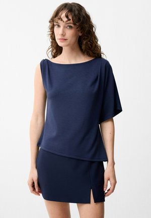 Kvinde med krøllet hår iført en mørkeblå asymmetrisk ærmeløs top og et matchende kort skørt med sideslidse, stående mod en ensfarvet baggrund.