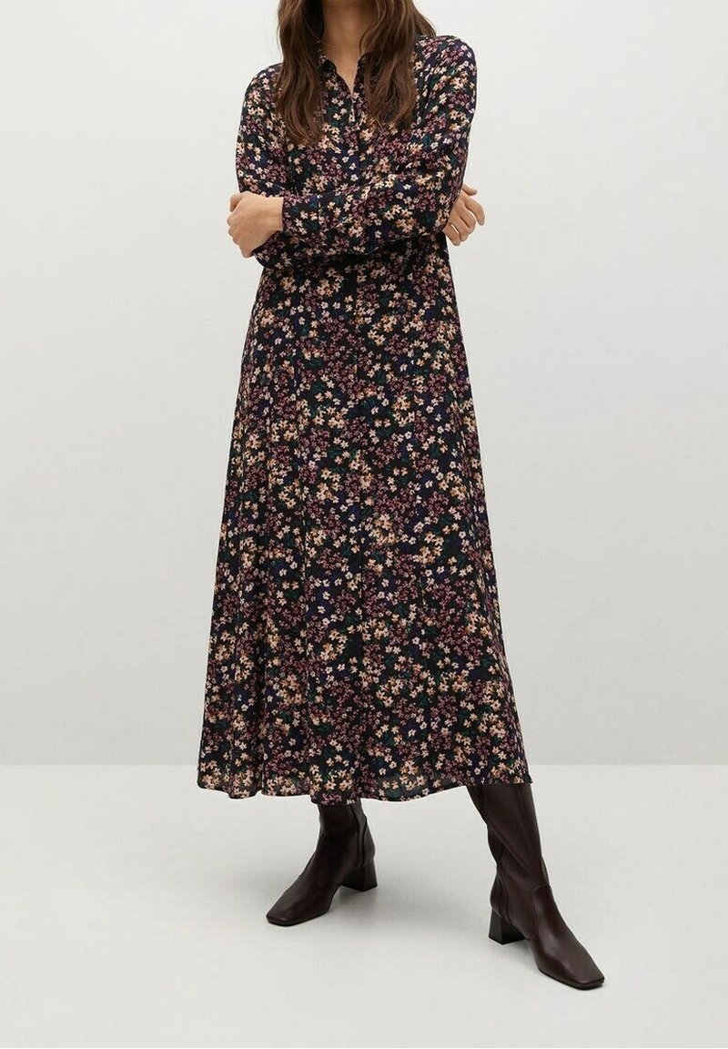 Femme portant une robe noire longue aux manches longues et longueur cheville avec un petit imprimé floral multicolore et des bottines marron foncé, debout, les bras croisés.
