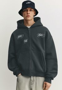 Fiatal férfi fekete kalapban és sötét zipzáras kapucnis pulóverben, amelyen absztrakt fehér szöveg található, világos, egyszínű háttér előtt áll.