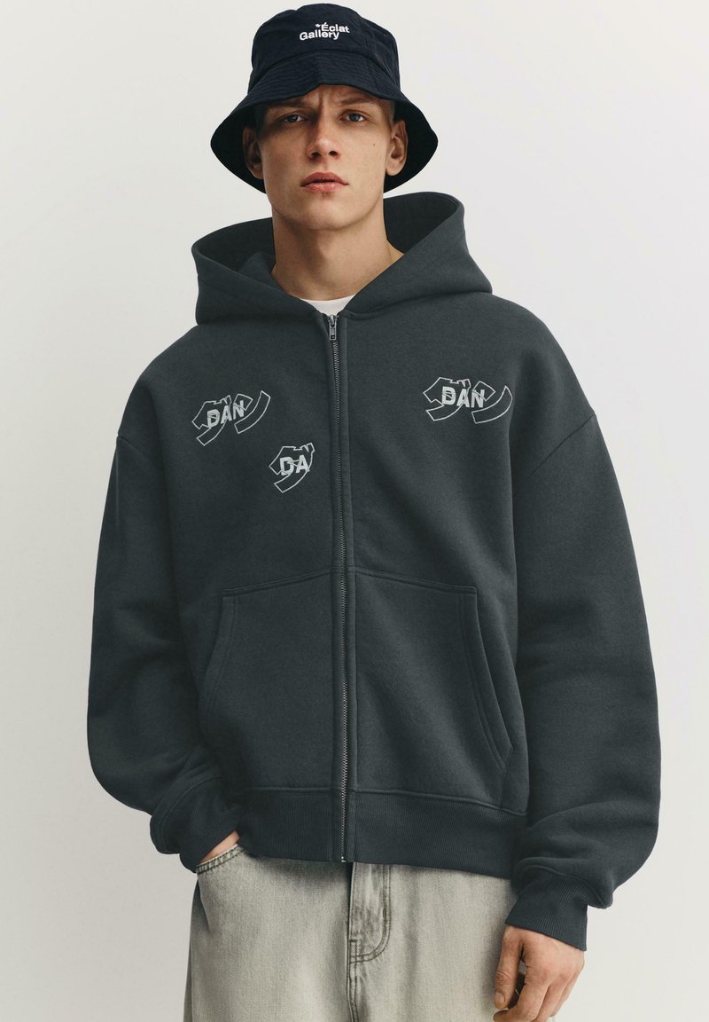 Fiatal férfi fekete kalapban és sötét zipzáras kapucnis pulóverben, amelyen absztrakt fehér szöveg található, világos, egyszínű háttér előtt áll.