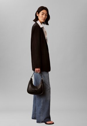 Femme portant un blazer noir, une chemise blanche, un jean large, tenant un sac à main CK noir, debout devant un fond gris uni.