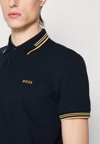 Polo shirt navy en coton, featuring a patte de boutonnage à trois boutons, logo doré, et accents rayés jaunes et noirs sur le col et les manches.
