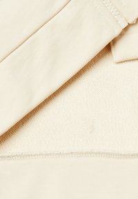 Tessuto beige con una texture liscia, dotato di una cucitura piatta, dettaglio con zip e uno strato sottostante morbido e intrecciato.