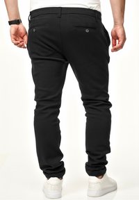 Schwarze Hosen mit tapered Fit, ausgestattet mit zwei Gesäßtaschen mit Knöpfen. Kombiniert mit weißen Sneakers, die eine glatte Textur zeigen.