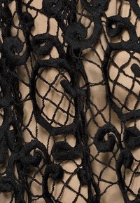 Superposition de dentelle noire brodée avec des motifs complexes en spirale et circulaires sur un fond de tissu beige clair, mettant en valeur la texture et la profondeur.