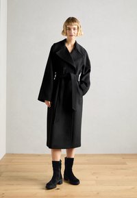 Dlouhý černý vlněný kabát s převislými klopami, dvouřadovým zapínáním na knoflíky a odpovídajícím páskem. Modelka má na sobě černé semišové kotníčkové boty.