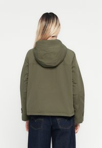 Une personne avec de longs cheveux blonds portant une jacket à capuche vert olive et un jean bleu foncé, faisant face à un fond blanc uni.