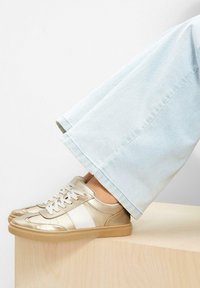 Sneaker metallici dorati con accenti bianchi, caratterizzati da una texture liscia, design con lacci e suola in gomma chiara, abbinati a denim chiaro.