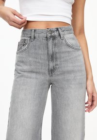 Jeans cu talie înaltă și picioare largi din denim gri deschis. Prezintă două buzunare frontale și închidere cu nasture. Texturat, cu o estompare subtilă.