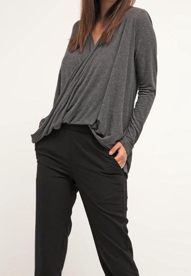 Femme portant un haut gris charbon à manches longues drapé, rentré dans un pantalon noir taille haute, mains dans les poches, debout devant un fond clair.