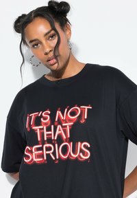 Zwart oversized t-shirt met opvallende rode neonletters en de tekst "HET IS NIET ZO SERIEUS." Korte mouwen en een ontspannen pasvorm.