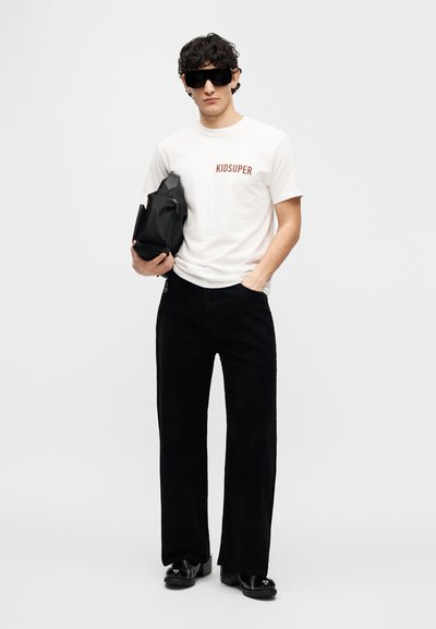 Λευκό κοντομάνικο Τ-Shirt με το «KIDSUPER» σε κόκκινο, συνδυασμένο με μαύρο παντελόνι κοτλέ και γυαλιστερά μαύρα παπούτσια. Το μοντέλο κρατάει μια μαύρη τσάντα.