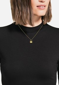 Kvinna med axellångt brunt hår som bär en svart topp och ett guldhalsband med en liten hänglåsberlock.