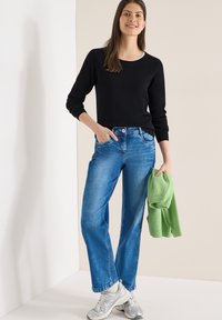 Femme debout portant un haut noir à manches longues, un jean bleu, des baskets blanches, tenant un pull vert plié dans une main.