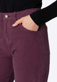 Lila Cord-Hosen mit flachem Front und Seitentasche. Merkmale sind Metallknöpfe und Nähdetails; weiche Textur mit vertikalen Rippen.
