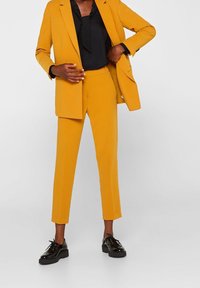 Senapsgul skräddarsydd kostym med en enkelknäppt kavaj och slim-fit byxor, kombinerad med en svart blus och blanka svarta skor.