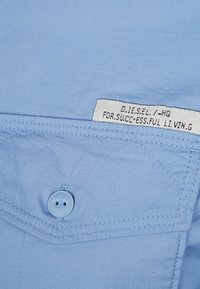 Vaaleansininen paita, jossa on etutasku napilla, näkyviä ommeltyylejä ja kangasmerkki, jossa on teksti.