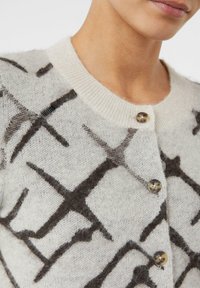 Sweter z jasnego dzianinowego materiału z ciemnym abstrakcyjnym wzorem. Żebrowany okrągły dekolt i dwa brązowe guziki z przodu.