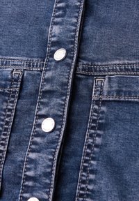 Gros plan sur une veste en jean bleu avec des boutons-pression en métal et des coutures doubles visibles autour des poches et de la patte de boutonnage.