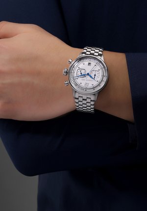Chronografur i rustfrit stål med sølvfarvet urskive, blå detaljer, datovisning og en sammenkoblet armbånd, båret på et håndled.