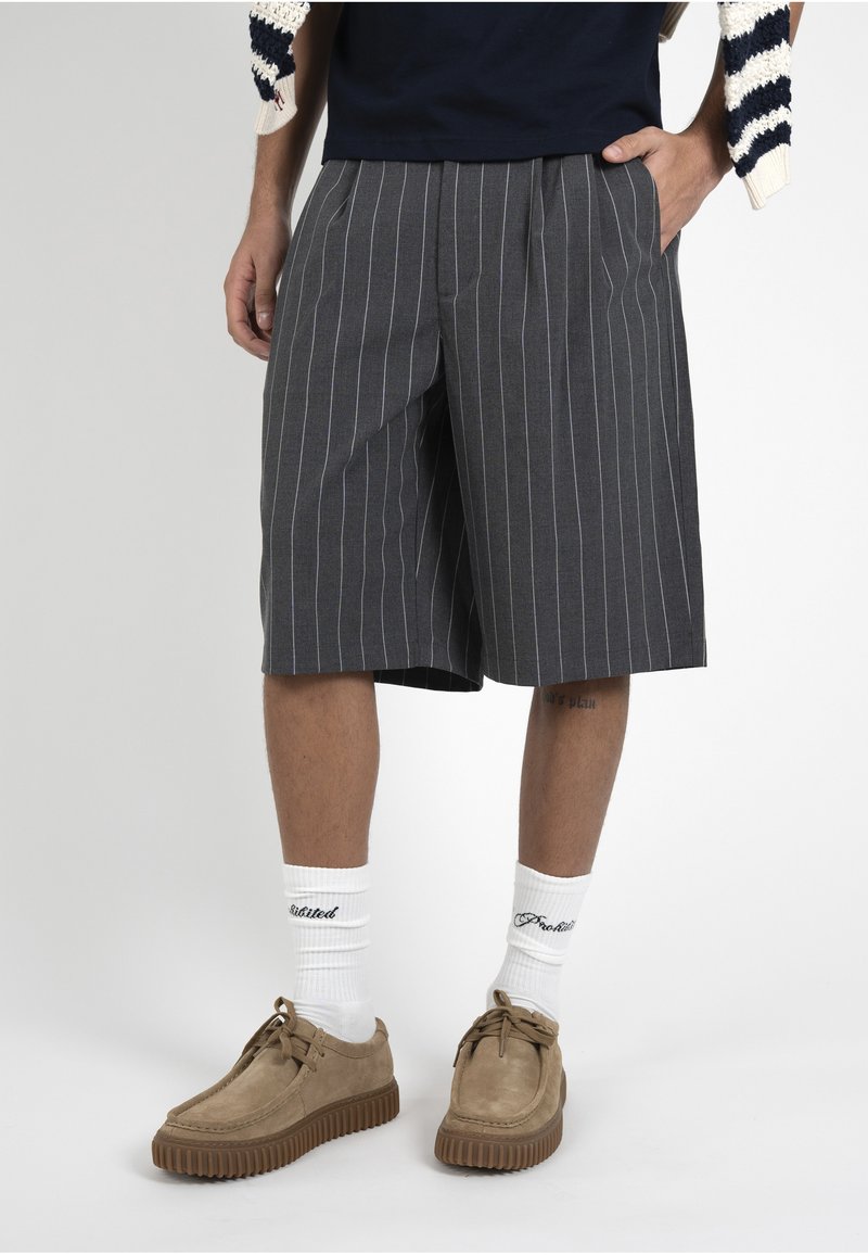 Personne portant un short gris foncé à fines rayures jusqu'aux genoux, des chaussettes blanches avec un texte noir, et des chaussures en daim beige à lacets avec des semelles épaisses.