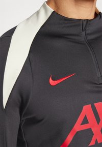 Koszulka treningowa Nike w czarnym kolorze z wysokim kołnierzem, białymi akcentami na ramionach oraz czerwonym logo swoosh.