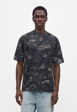 Jeune homme portant une chemise noire à manches courtes à motif floral et un jean bleu clair sur fond blanc.