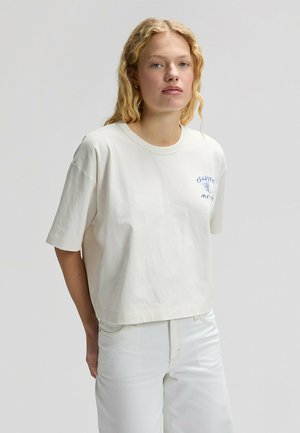 Blonde vrouw die een off-white korte mouw cropped t-shirt met blauwe tekst op de borst en off-white hoge taille spijkerbroek draagt, staand.