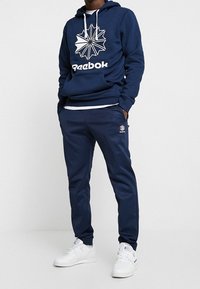 Felpa blu navy con logo Reebok bianco e grafica geometrica. Completa con pantaloni jogger blu navy coordinati e sneakers bianche.