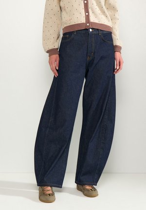 Vrouw draagt donkere jeans met wijde pijpen, beige gebreid vest met knopen en lichtbruine platte schoenen, staand tegen een lichte achtergrond.