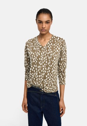 Kobieta w brązowym swetrze z dekoltem w serek z białymi nieregularnymi kropkami i ciemnoniebieskich jeansach z wysokim stanem, stojąca na tle jednolitego jasnego tła.