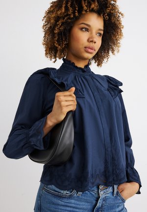 Nuori nainen, jolla on kiharat hiukset, pukeutuneena tummansiniseen röyhelöpaitaan ja farkkuihin, kantaa pientä mustaa nahkaista olkalaukkua.